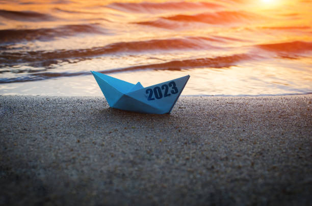 istock Blue paper boat with inscription 2023, on sandy shore beach against sunset. 1500251685