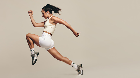 Sportswoman jumping forward, running in a vigorous studio workout. Wearing sportswear, she combines strength training with cardio exercises to improve fitness and performance. Female athlete pushing herself to new limits.