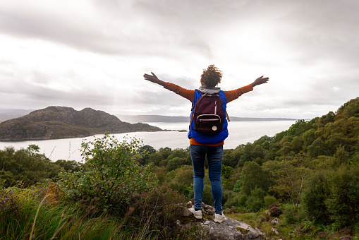 Rear view of a mature woman hiking up the mountain landscape surrounding Loch Torridon in the Northwest Highlands, Scotland. She has stopped to stand on a rock, she has her arms outstretched and she is feeling at one with nature.