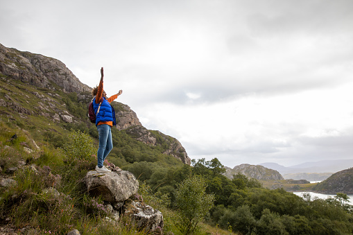 Side view of a mature woman hiking up the mountain landscape surrounding Loch Torridon in the Northwest Highlands, Scotland. She has stopped to stand on a rock, she has her arms outstretched and she is feeling at one with nature.
