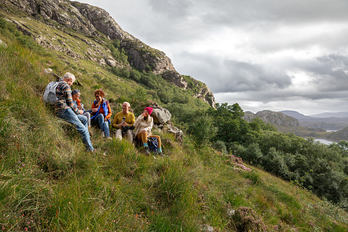 Wide view of a small group of mature and senior friends sitting down and taking a break from their hike up the mountain landscape surrounding Loch Torridon in the Northwest Highlands, Scotland to sit, relax and chat while they enjoy eating their lunch.