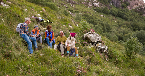 A wide view of a small group of mature and senior friends sitting down and taking a break from their hike up the mountain landscape surrounding Loch Torridon in the Northwest Highlands, Scotland to sit, relax and chat while they enjoy eating their lunch.