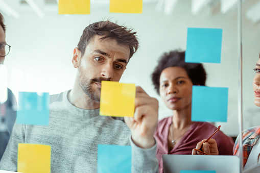 Through glass view of male and female software developer executives in casual wear using post it notes while sharing ides at start-up company