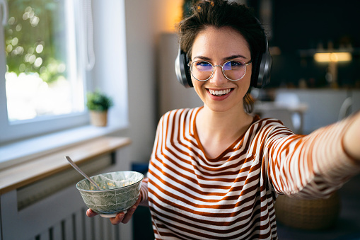Young Caucasian woman with wireless headphones taking selfie while holding an oatmeal breakfast in a bowl.