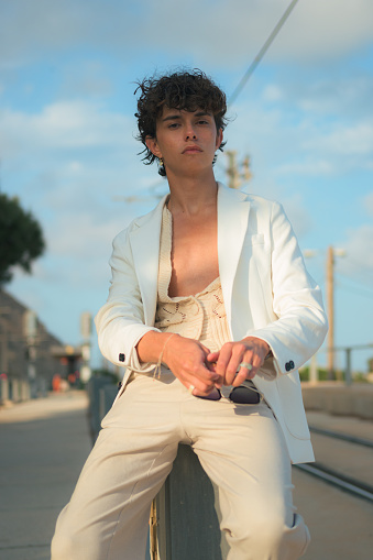 Young gay man in white clothes is posing on the street at sunset
