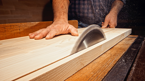 Close-up of male carpenter's hands cutting wooden plank with table saw in workshop.