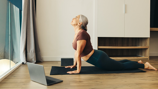 Senior woman practices upward facing dog pose on her fitness mat, feeling the strength and flexibility in her body. Mature woman finding focus and balance in her home yoga routine.