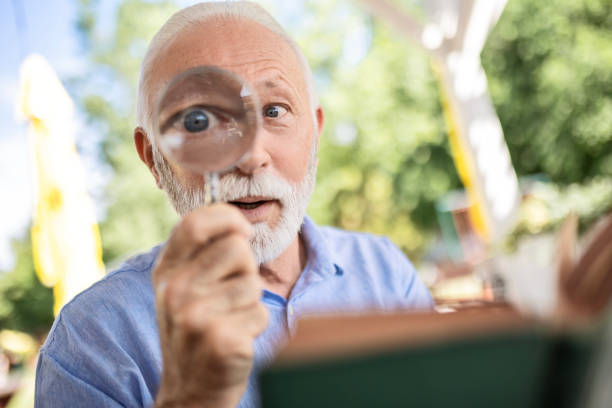 uomo anziano con la lente d'ingrandimento nel libro di lettura della mano - lente bulbo oculare foto e immagini stock