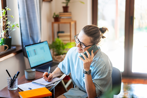 Beautiful  young adult woman  businesswoman  freelancer speaking on smart phone  working at home using laptop