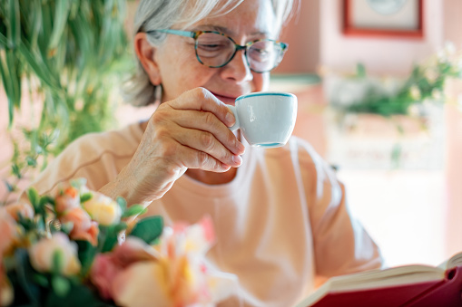 Blurred senior woman sitting at cafe table reading a book or studying enjoying an espresso coffee. Elderly lady holding a white coffee cup