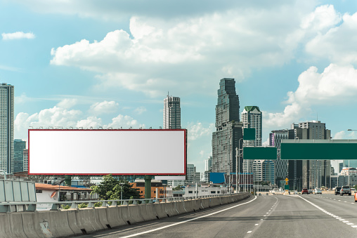 A large billboard is on a city street with a yellow sign in front of it. The city is bustling with activity and the sky is cloudy. Bangkok, Thailand.