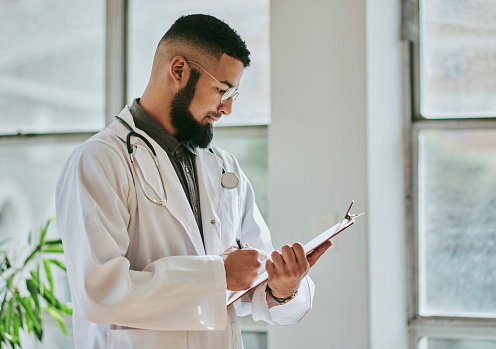A side view of a  young male doctor standing while looking down at a document on a clipboard, reading and writing on the document with copy space, stock photo