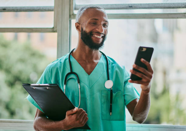 若い黒人男性看護師の正面図は、ビデオ通話で携帯電話を持って立っています。ストックフォト - male nurse medical student healthcare and medicine nursing assistant ストックフォトと画像