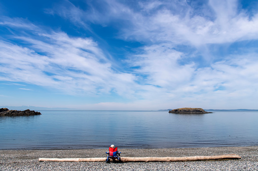 View from Rosario Beach in Deception Pass State Park, WA, USA towards the water of Rosaria Strait with lone man in chair facing water against piece of driftwood and blue sky with white feather clouds