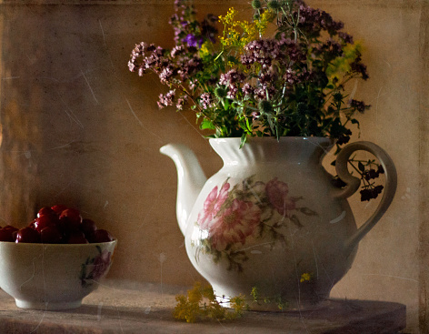 Still life with wildflowers in a vintage teapot. Nearby on a wooden table are cherries in a ceramic bowl. Image stylization for old photos with scratches.