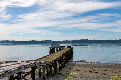 Panoramic view over the water of Penn Cove near Coupeville, WA, USA with a wooden fishing dock and building extended in the water against a blue sky with white feather clouds