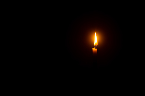 A single burning candle flame or light is glowing on an orange candle on black or dark background on table in church for Christmas, funeral or memorial service with copy space.