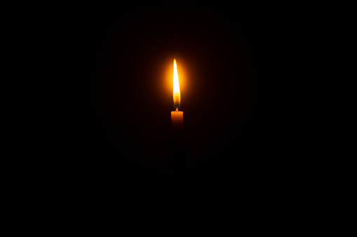 A single burning candle flame or light is glowing on an orange candle on black or dark background on table in church for Christmas, funeral or memorial service with copy space.