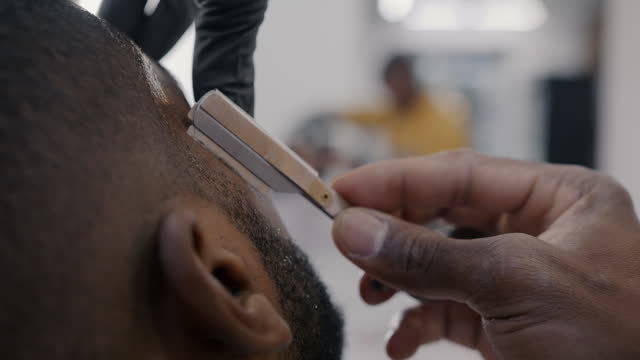 Close-up of hand with sharp blade shaving beard on male face while barber taking care of client in barbershop