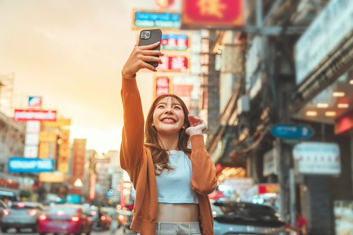 Happy young Asian tourist woman selfie by mobile smartphone, Female traveller taking photo of Chinatown street food market during sunset in Bangkok, Thailand, Summer journey trip concept.