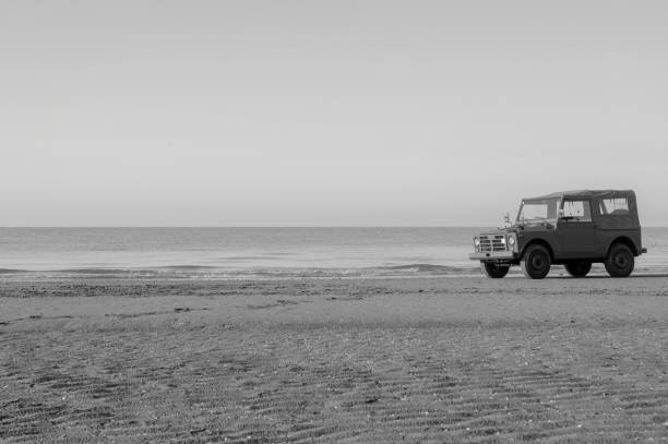 a vintage italian 4x4 jeep paked near the sea in an empty beach in summer at sunset. Young woman pose in fashionable clothes on the sand beach seat over a vintage jeep.. Beautiful sea landscape on background. Brunette model, tall, tan and slim. Summer, sunset. Black and white picture. sequence dress stock pictures, royalty-free photos & images