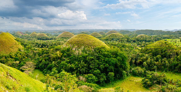 A panoramic view of the famous Chocolate Hills on Bohol Island, Philippines. The landscape is dominated by the presence of the Chocolate Hills, which are a series of conical-shaped hills covered in lush tropical plants. The hills extend as far as the eye can see, creating a captivating sight. The sky above is blue with scattered clouds, adding depth to the overall scene. This photograph beautifully captures the natural beauty of Bohol Island, specifically highlighting the unique geological formations known as the Chocolate Hills.