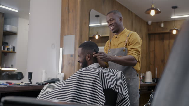 Cheerful barber in apron working with client shaving hair indoors in modern barbershop