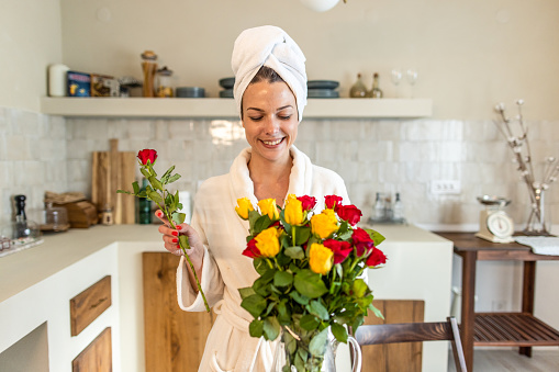Happy young woman standing over her table and arranging roses in a glass vase to decorate her home with.