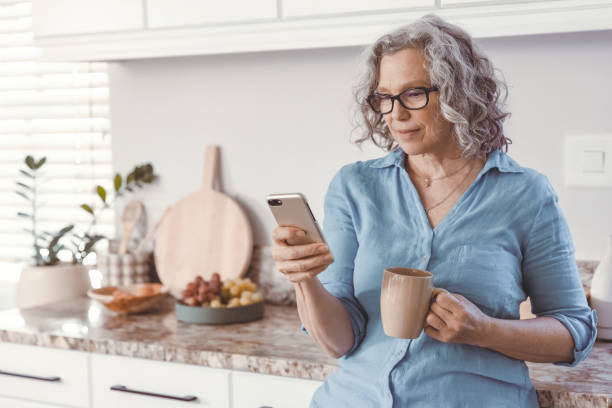 femme âgée debout dans la cuisine avec son téléphone - women telephone senior adult on the phone photos et images de collection