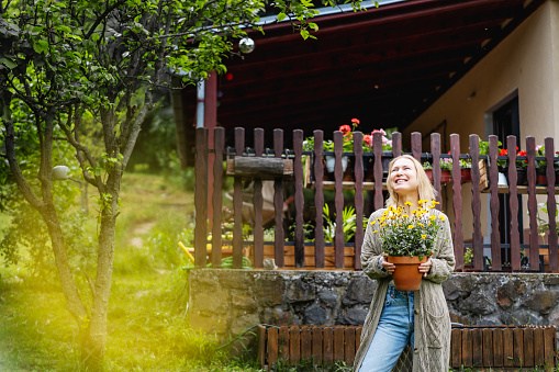 Happy middle-aged blonde woman with a pot of flowers in her hands on the background of a country house. Summer lifestyle