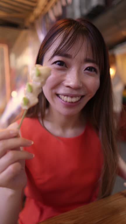 Business woman in orange dress live streaming in Izakaya bar after work, eating ‘Yakitori’ Japanese chicken skewer