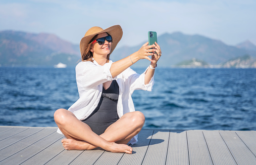 Woman in a hat sunglasses and a white shirt sitting on a pier on the seashore making a selfie on a smartphone during a summer vacation travel