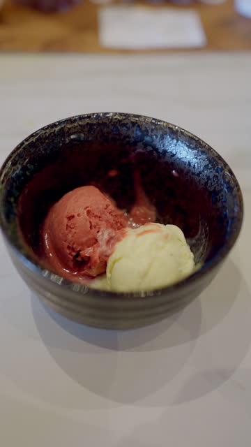 Scoops of raspberry and vanilla ice cream in the black bowl, one human hand spooning the red one a bit to taste with a small spoon