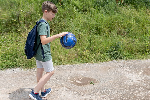 A boy with backpack and football ball is going home after football training.  A boy walking along the dirt road. He is playing with ball.