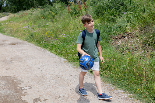 A boy with backpack and football ball is going home after football training.  A boy walking along the dirt road. He is playing with ball.