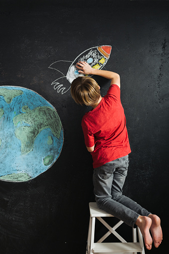 Rear view of  boy drawing rocket and stars next to a planet earth chalk drawing on blackboard wall