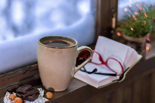 Winter cozy still life with cup of coffee, book, Eyewear, and Christmas tree. Snowdrifts outside the window. Hygge.