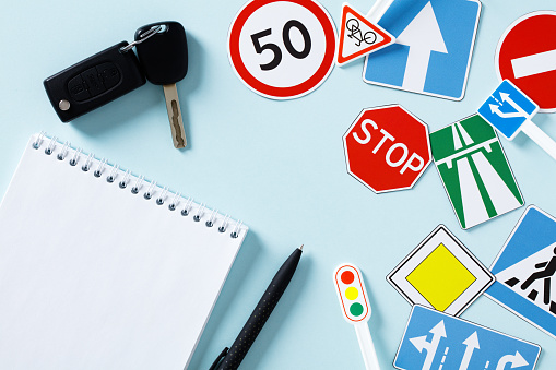 Flat lay paper notebook, car keys, road sign on blue background. Driving school concept.
