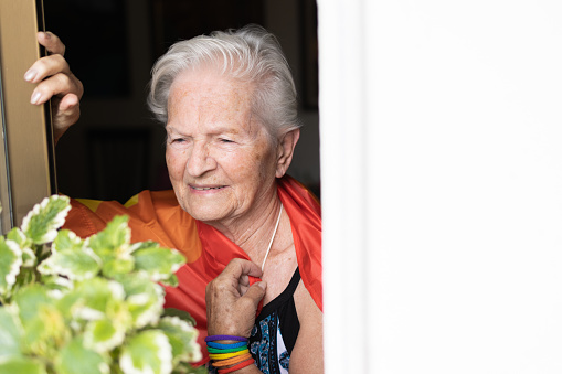 elderly caucasian lesbian woman with rainbow flag and wristband looking out the window at the gay pride day parade. lbgt and homosexual concept.cheerful smile