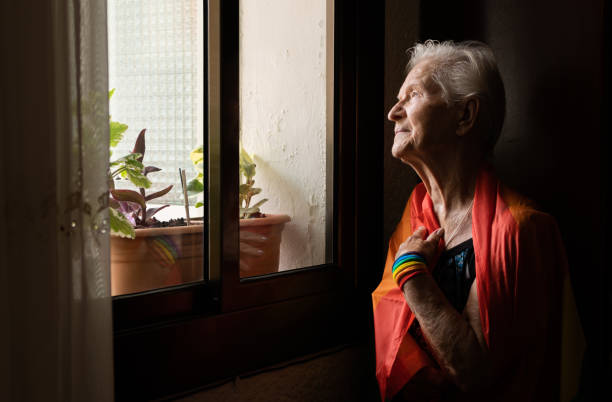 donna lesbica caucasica anziana con bandiera arcobaleno e braccialetto che guarda fuori dalla finestra alla parata del gay pride day. lbgt e concetto omosessuale. - gay pride wristband rainbow lgbt foto e immagini stock