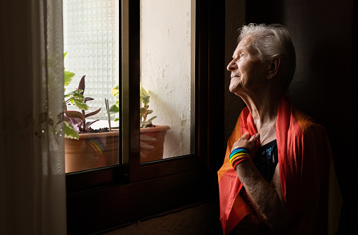 elderly caucasian lesbian woman with rainbow flag and wristband looking out the window at the gay pride day parade. lbgt and homosexual concept.
