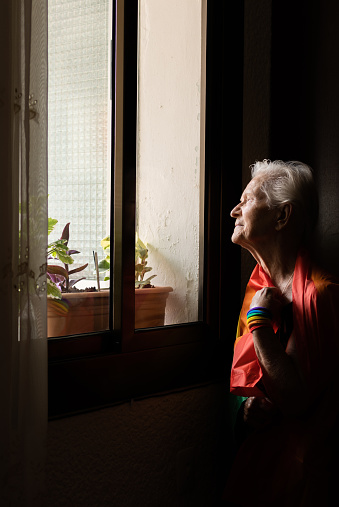 elderly caucasian lesbian woman with rainbow flag and wristband looking out the window at the gay pride day parade. lbgt and homosexual concept.