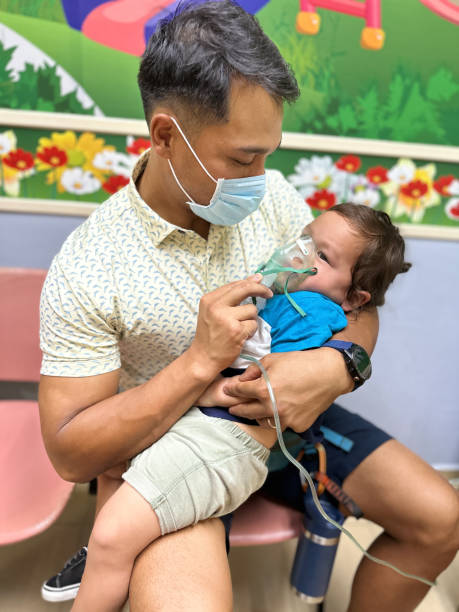 Father helping sick son use nebulizer while at a doctor's appointment Filipino father holding his Eurasian toddler son in his lap and helping the child use a nebulizer while at the hospital due to RSV and pneumonia. pediatric nebulizer mask stock pictures, royalty-free photos & images