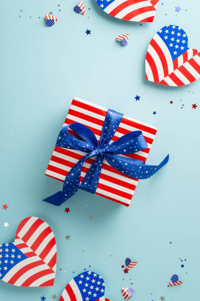 Happy 4th of July celebration. Vertical overhead shot of patriotic-themed decorations including American flag-patterned hearts, confetti, and big gift box in thematic wrapping, on a soft blue backdrop feliz celebración del 4 de julio. toma aérea vertical de decoraciones de temática patriótica que incluyen corazones estampados con la bandera estadounidense, confeti y una gran caja de regalo en envoltura temática, sobre un fondo azul suave
