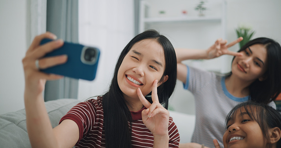 Three Asian Siblings women gen z in great mood takes selfie in living room. she holding smartphone and taking picture of herself and sister. Happy woman portrait concept