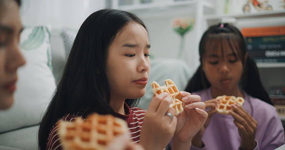 Selective focus, Three Asian Siblings women gen z with party waffles in living room She enjoy eating and fun. During rest time, young women eat waffles and chatted hilariously.