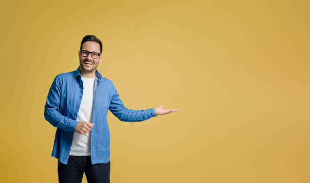 Portrait of happy salesman showing something on palm while standing against yellow background Portrait of happy salesman showing something on palm while standing against yellow background spokesmodel stock pictures, royalty-free photos & images