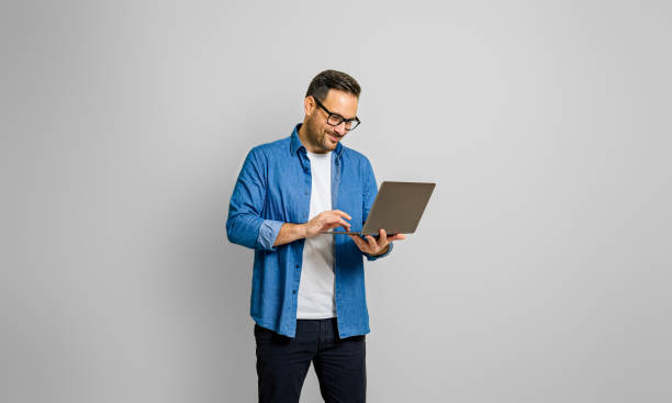 selbstbewusster männlicher unternehmer analysiert bericht über laptop, während er vor weißem hintergrund steht - isolated cheerful businessman smiling stock-fotos und bilder