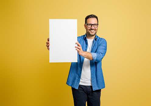 Happy male marketing manager showing blank white poster for advertising against yellow background
