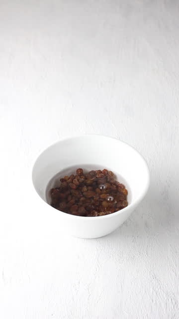 Raisins are poured into a white bowl and filled with water for soaking and rinsing on a gray textured background